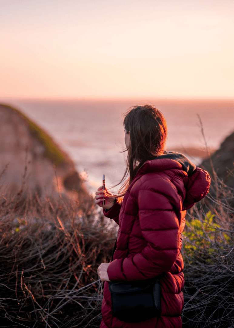 A woman wearing a red jacket holding her weed vape pen from Xzibit's West Coast Cannabis while standing on a cliff overlooking one of LA's beaches at sunset.